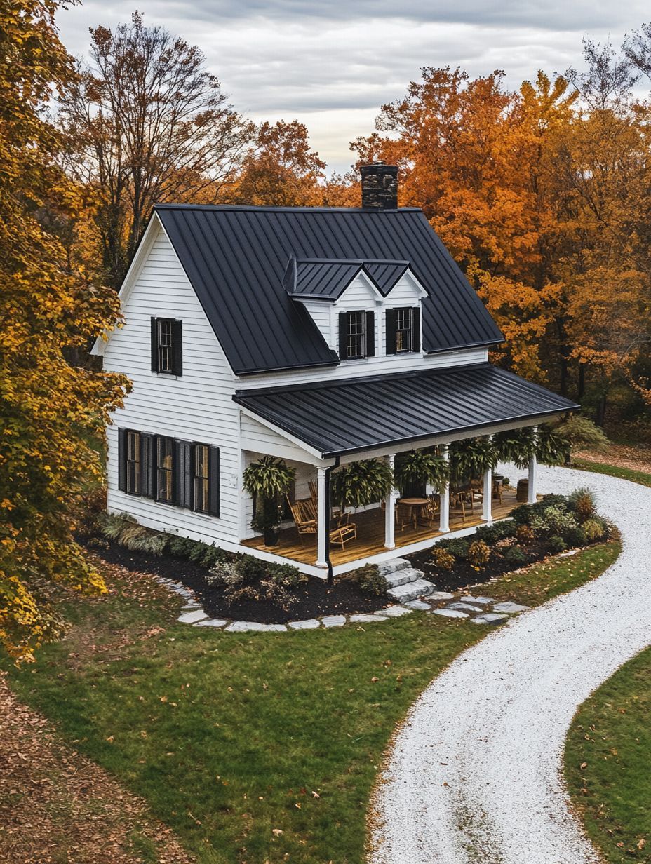 A white two-story house with a black metal roof and a wraparound porch sits next to a curved gravel driveway, surrounded by autumn trees with orange leaves.