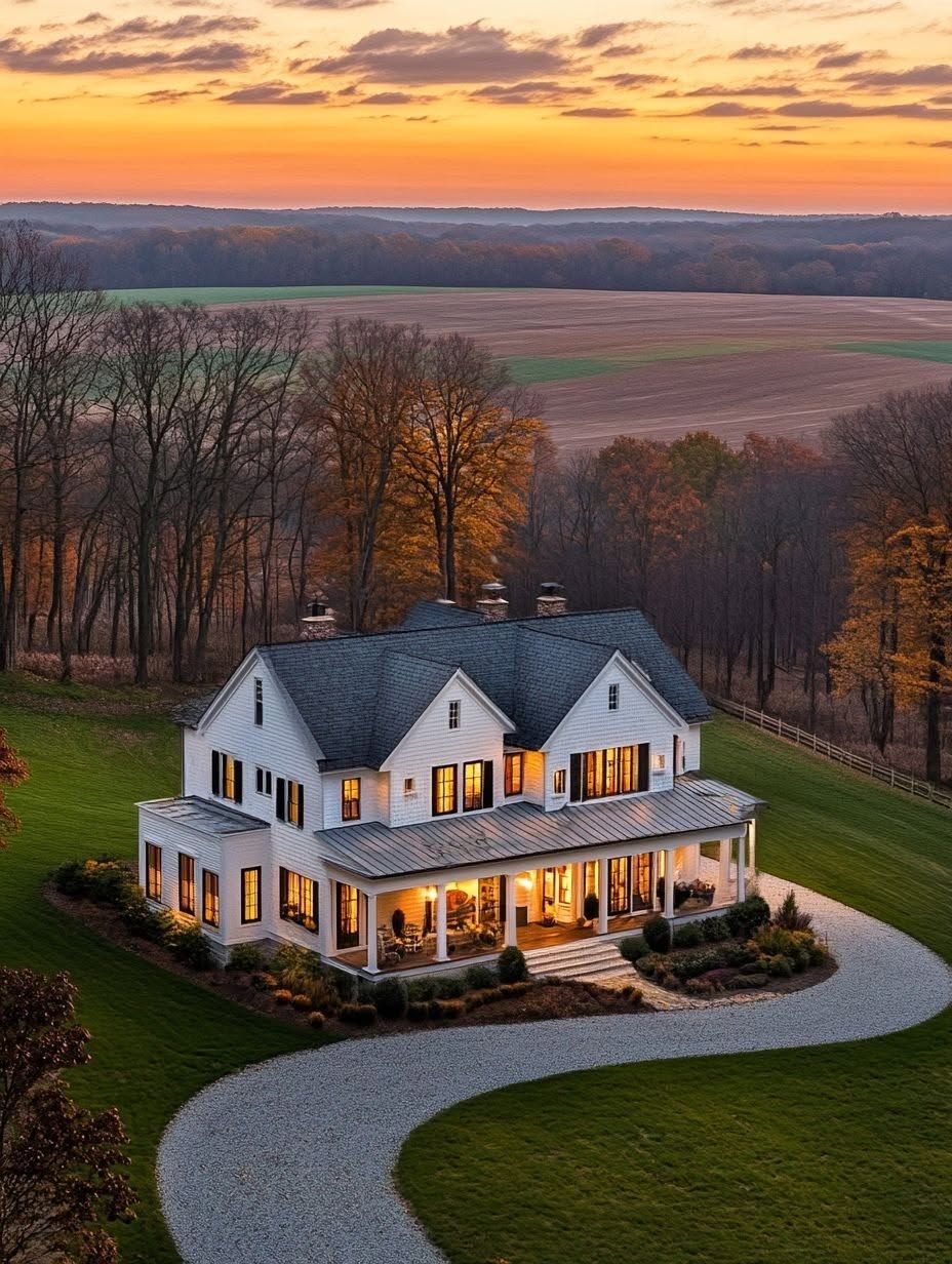 Two-story white house with a wraparound porch and lit windows, surrounded by a lawn and trees, with fields and a sunset sky in the background.