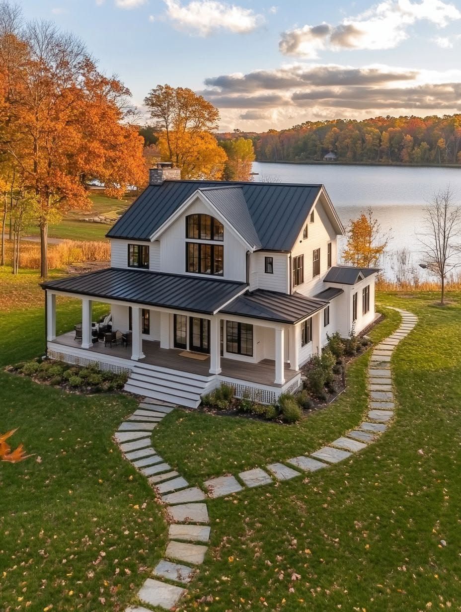 A modern white farmhouse with black roof sits by a lake, surrounded by autumn trees and a curved stone pathway on a green lawn.