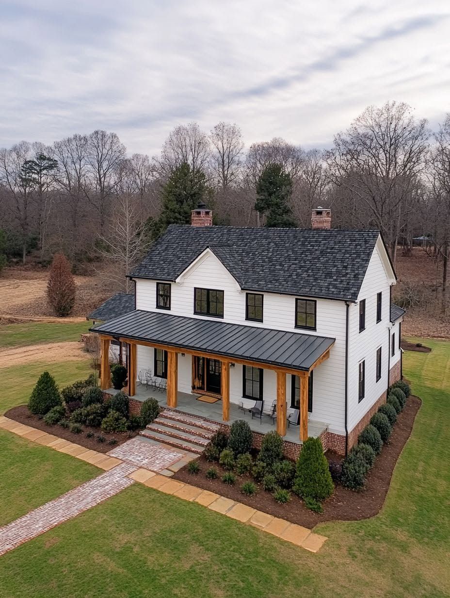 Two-story white house with black roof, wooden pillars on a front porch, surrounded by trimmed bushes, lawn, and a brick walkway, with trees and cloudy sky in the background.